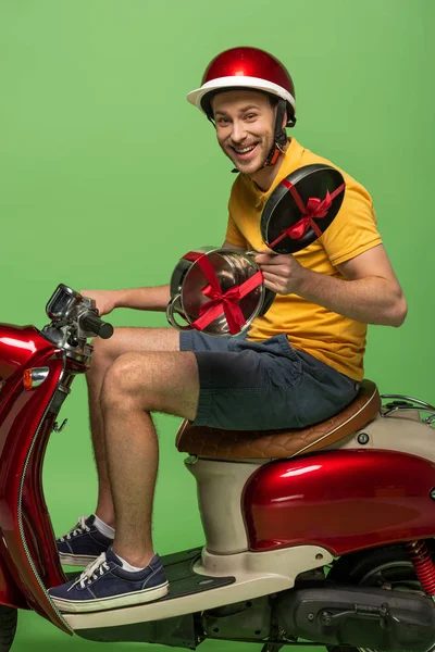 Side view of happy delivery man in yellow uniform holding frying pan with bow on scooter isolated on green — Stock Photo