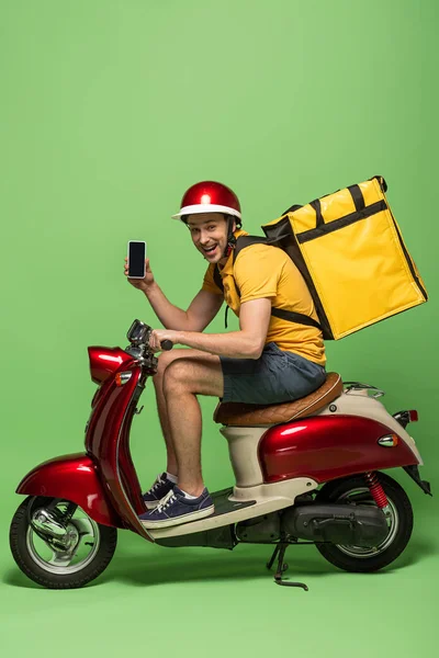 Side view of happy delivery man in yellow uniform with backpack showing smartphone with blank screen on scooter on green — Stock Photo