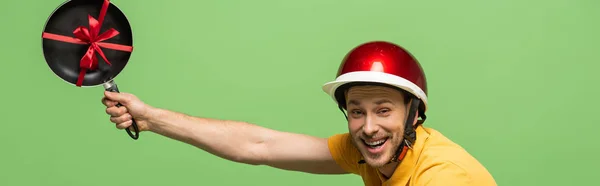 Side view of happy delivery man in yellow uniform holding frying pan with bow isolated on green, panoramic shot — Stock Photo