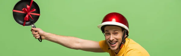 Visão lateral do homem de entrega feliz em amarelo uniforme segurando frigideira com arco isolado em verde, tiro panorâmico — Fotografia de Stock