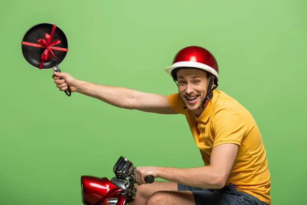 Visão lateral do homem de entrega feliz em amarelo uniforme segurando frigideira com arco em scooter isolado no verde — Fotografia de Stock