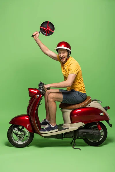 Visão lateral do homem de entrega feliz em amarelo uniforme segurando frigideira com arco em scooter no verde — Fotografia de Stock