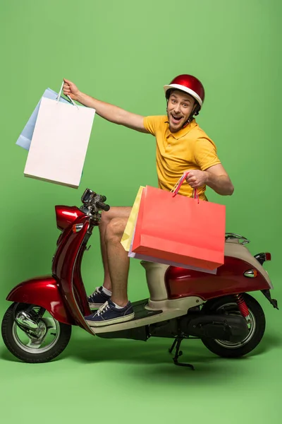 Вид сбоку счастливого доставщика в желтой форме и шлеме, держащего сумки с покупками на скутере на зеленом — стоковое фото