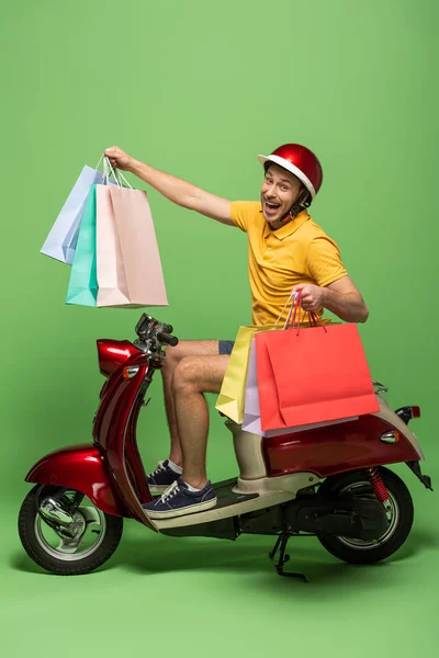 Seitenansicht eines glücklichen Zustellers in gelber Uniform und Helm, der Einkaufstüten auf einem Roller auf Grün hält — Stockfoto