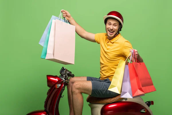 Вид сбоку улыбающегося доставщика в желтой форме и шлеме, держащего сумки с покупками на скутере на зеленом — стоковое фото