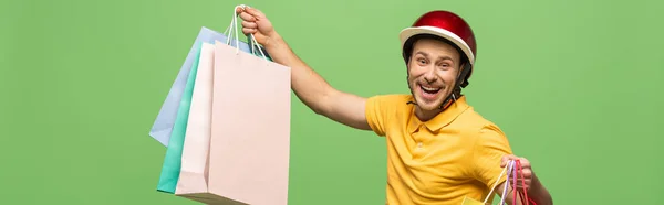 Repartidor sonriente en uniforme amarillo y casco sosteniendo bolsas aisladas en verde, tiro panorámico - foto de stock