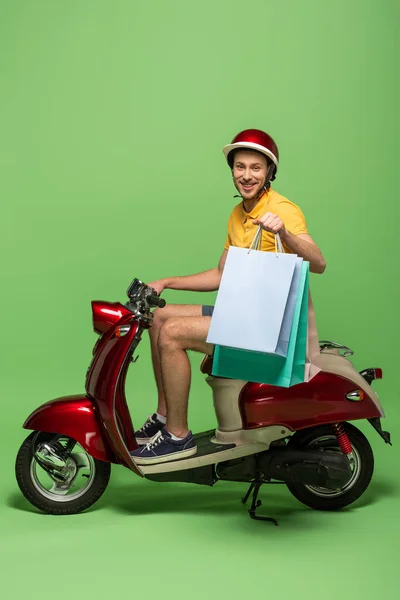 Vista lateral del repartidor sonriente en uniforme amarillo y casco sosteniendo bolsas de compras en scooter en verde — Stock Photo