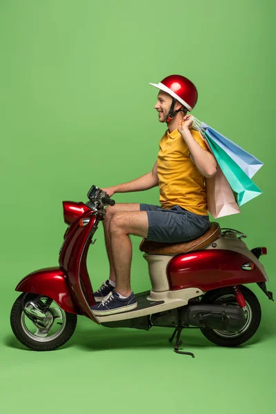 Vista laterale di uomo consegna sorridente in uniforme gialla e casco che tiene borse della spesa su scooter su verde — Foto stock