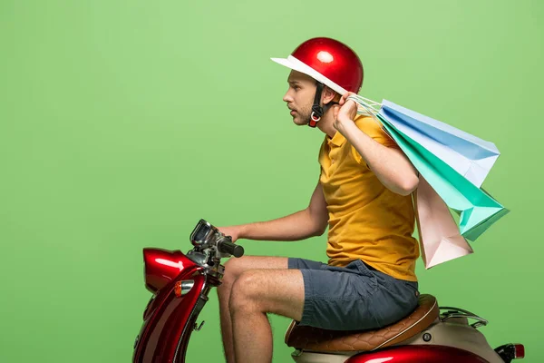 Vista lateral del repartidor hombre en uniforme amarillo y casco sosteniendo bolsas de compras en scooter aislado en verde — Stock Photo