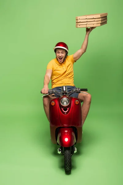 Excited delivery man in yellow uniform with open mouth on scooter delivering pizza on green — Stock Photo