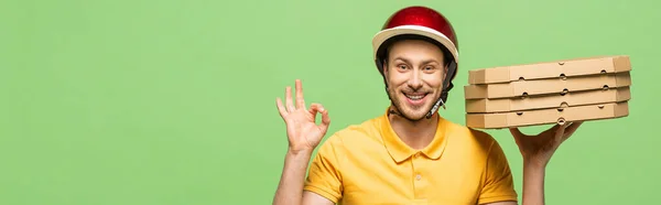 Lächelnder Lieferant in gelber Uniform, der Pizza ausliefert und auf grünem, panoramischem Foto ein Ok-Zeichen zeigt — Stockfoto