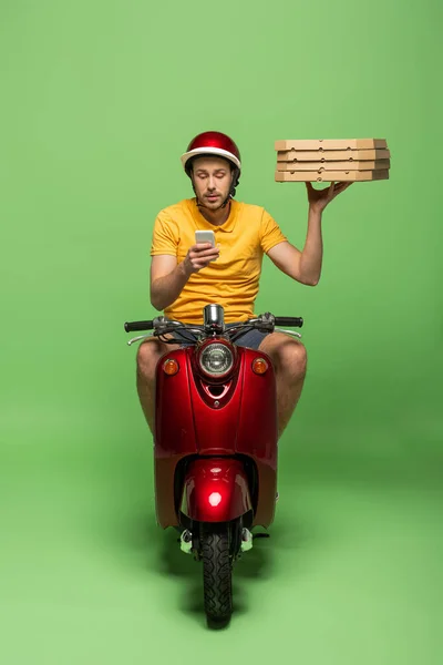 Homem de entrega em uniforme amarelo em scooter entregando pizza e usando smartphone no verde — Fotografia de Stock