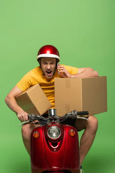 Сердитий чоловік доставки в жовтій формі на скутері з коробками, що говорять на смартфоні ізольовані на зеленому — стокове фото