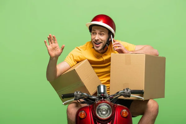 Happy delivery man in yellow uniform on scooter with boxes talking on smartphone and waving hand isolated on green — Stock Photo