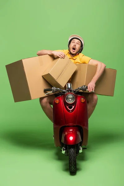 Livreur en uniforme jaune avec bouche ouverte sur scooter avec boîtes sur vert — Photo de stock