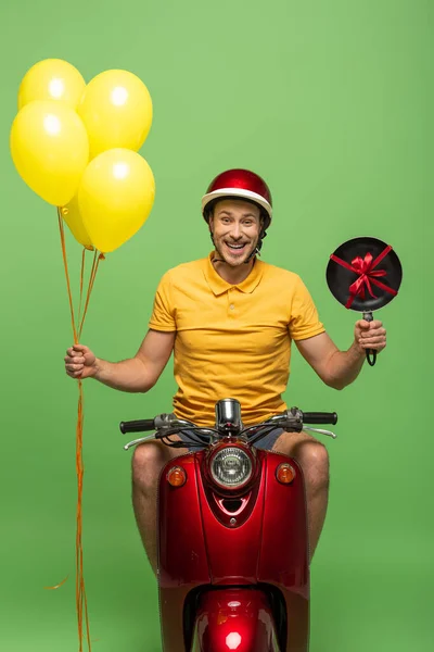 Hombre de entrega feliz en uniforme amarillo en scooter con sartén y globos aislados en verde - foto de stock