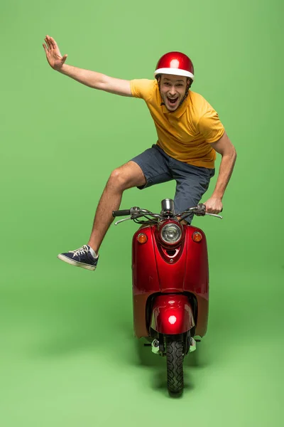 Hombre entrega feliz en uniforme amarillo y casco haciendo truco en scooter en verde - foto de stock