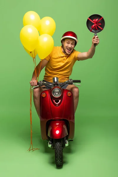 Щасливий чоловік доставки в жовтій формі на скутері зі сковородою і повітряними кулями на зеленому — стокове фото