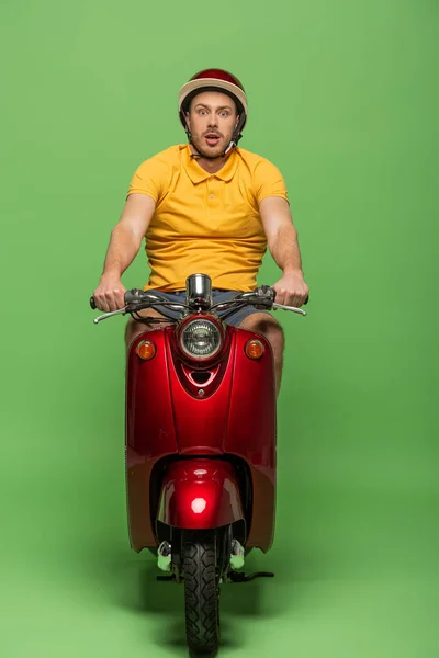 Chocado repartidor hombre en uniforme amarillo y casco en scooter en verde - foto de stock