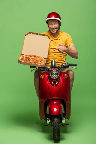 Lächelnder Zusteller in gelber Uniform auf Roller zeigt mit Finger auf Pizza auf Grün — Stockfoto