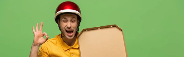 Winking delivery man in yellow uniform with pizza showing ok sign isolated on green, panoramic shot — Stock Photo