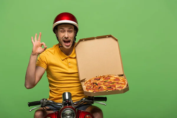 Winking delivery man in yellow uniform on scooter with pizza showing ok sign isolated on green — Stock Photo