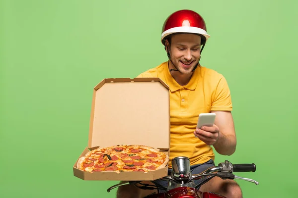 Smiling delivery man in yellow uniform on scooter with pizza using smartphone isolated on green — Stock Photo