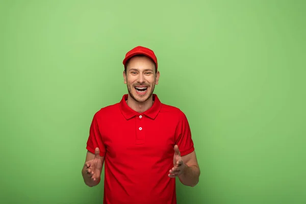 Щасливий чоловік доставки в червоній уніформі жестикулює на зеленому — стокове фото