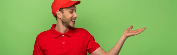 Homem de entrega feliz em uniforme vermelho apontando com a mão de lado isolado em verde, tiro panorâmico — Fotografia de Stock