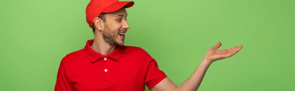 Feliz repartidor en uniforme rojo señalando con la mano a un lado aislado en verde, tiro panorámico - foto de stock