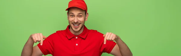 Smiling delivery man in red uniform pointing with finger down isolated on green, panoramic shot — Stock Photo