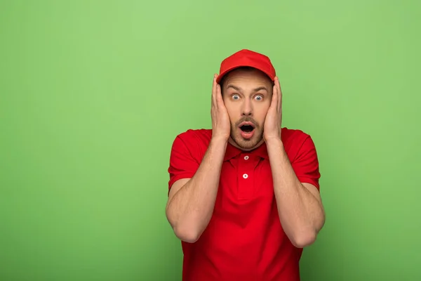 Scared delivery man in red uniform with open mouth touching face on green — Stock Photo