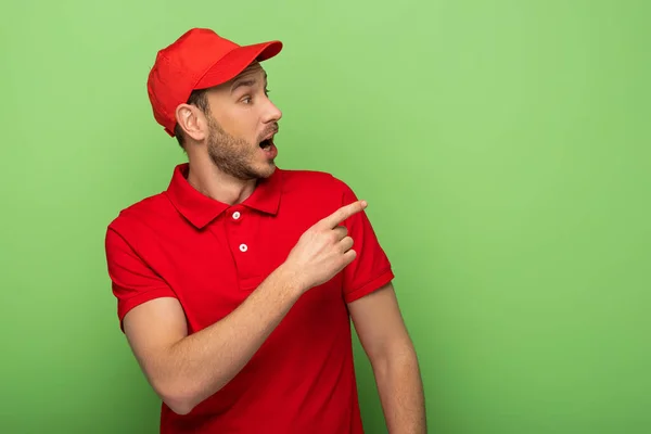 Schockierter Zusteller in roter Uniform zeigt mit Finger auf Grün — Stockfoto