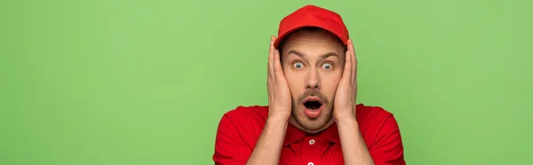 Scared delivery man in red uniform with open mouth touching face isolated on green, panoramic shot — Stock Photo