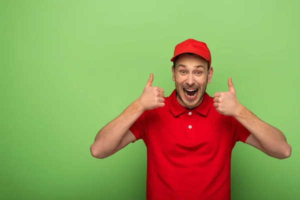 Eccitato uomo di consegna in uniforme rossa mostrando pollici in su sul verde — Foto stock