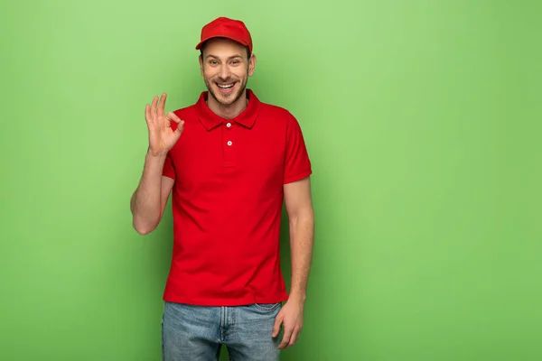 Усміхнений чоловік доставки в червоній формі, що показує знак ОК на зеленому фоні — стокове фото