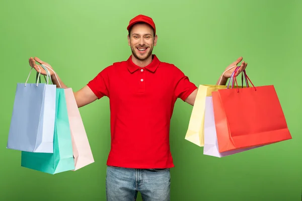 Hombre entrega feliz en uniforme rojo sosteniendo bolsas de compras en verde - foto de stock