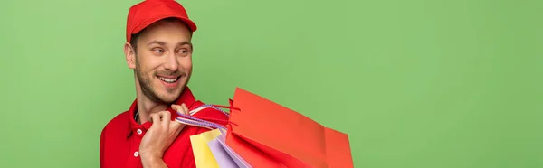 Щасливий чоловік доставки в червоній формі тримає сумки для покупок ізольовані на зеленому, панорамний постріл — стокове фото