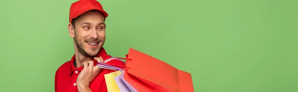 Feliz repartidor en uniforme rojo sosteniendo bolsas aisladas en verde, tiro panorámico - foto de stock