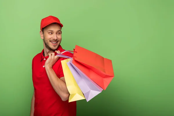 Hombre entrega feliz en uniforme rojo sosteniendo bolsas de compras en verde — Stock Photo