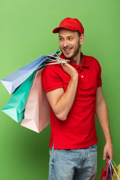 Hombre entrega feliz en uniforme rojo sosteniendo bolsas de compras en verde — Stock Photo
