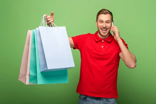 Hombre feliz sosteniendo bolsas de compras y hablando en el teléfono inteligente en verde - foto de stock