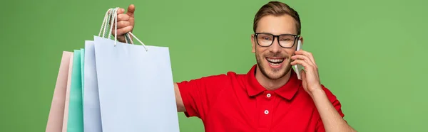 Felice uomo in occhiali in possesso di borse della spesa e parlando su smartphone isolato su verde, colpo panoramico — Foto stock