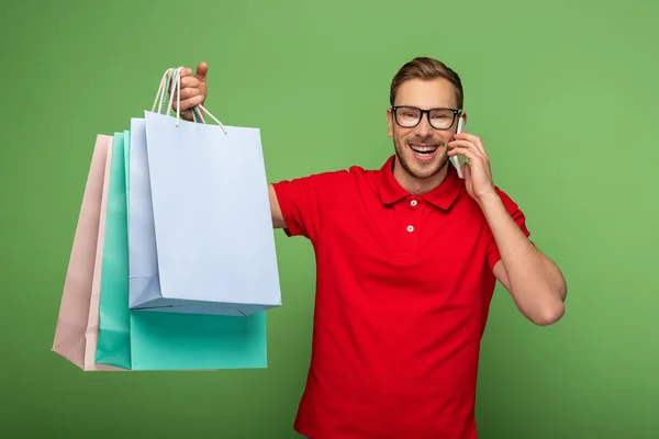 Happy man in eyeglasses holding shopping bags and talking on smartphone on green — Stock Photo