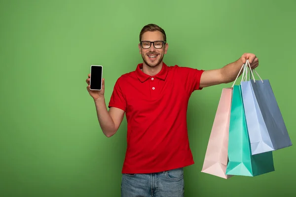 Happy man in eyeglasses holding shopping bags and smartphone on green — Stock Photo