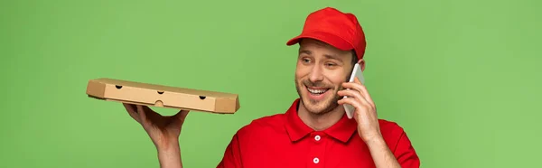 Felice uomo di consegna in uniforme rossa che tiene la scatola della pizza e parla su smartphone isolato sul verde, colpo panoramico — Foto stock