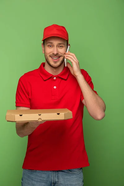 Hombre de entrega feliz en uniforme rojo sosteniendo la caja de pizza y hablando en el teléfono inteligente en verde - foto de stock