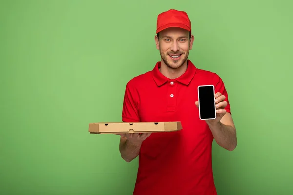 Repartidor sonriente en uniforme rojo sosteniendo la caja de pizza y el teléfono inteligente en verde - foto de stock