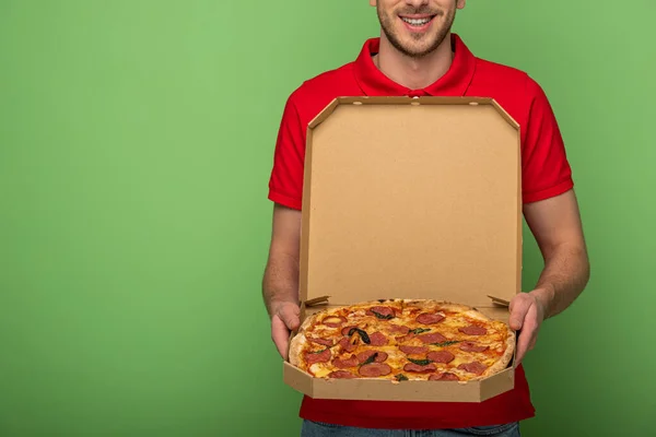 Corte vista de homem de entrega sorridente em vermelho uniforme segurando caixa de pizza no verde — Fotografia de Stock