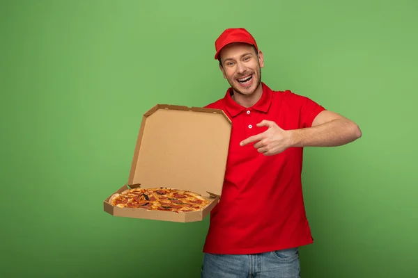 Hombre de entrega feliz en uniforme rojo apuntando con el dedo a la caja de pizza en verde - foto de stock